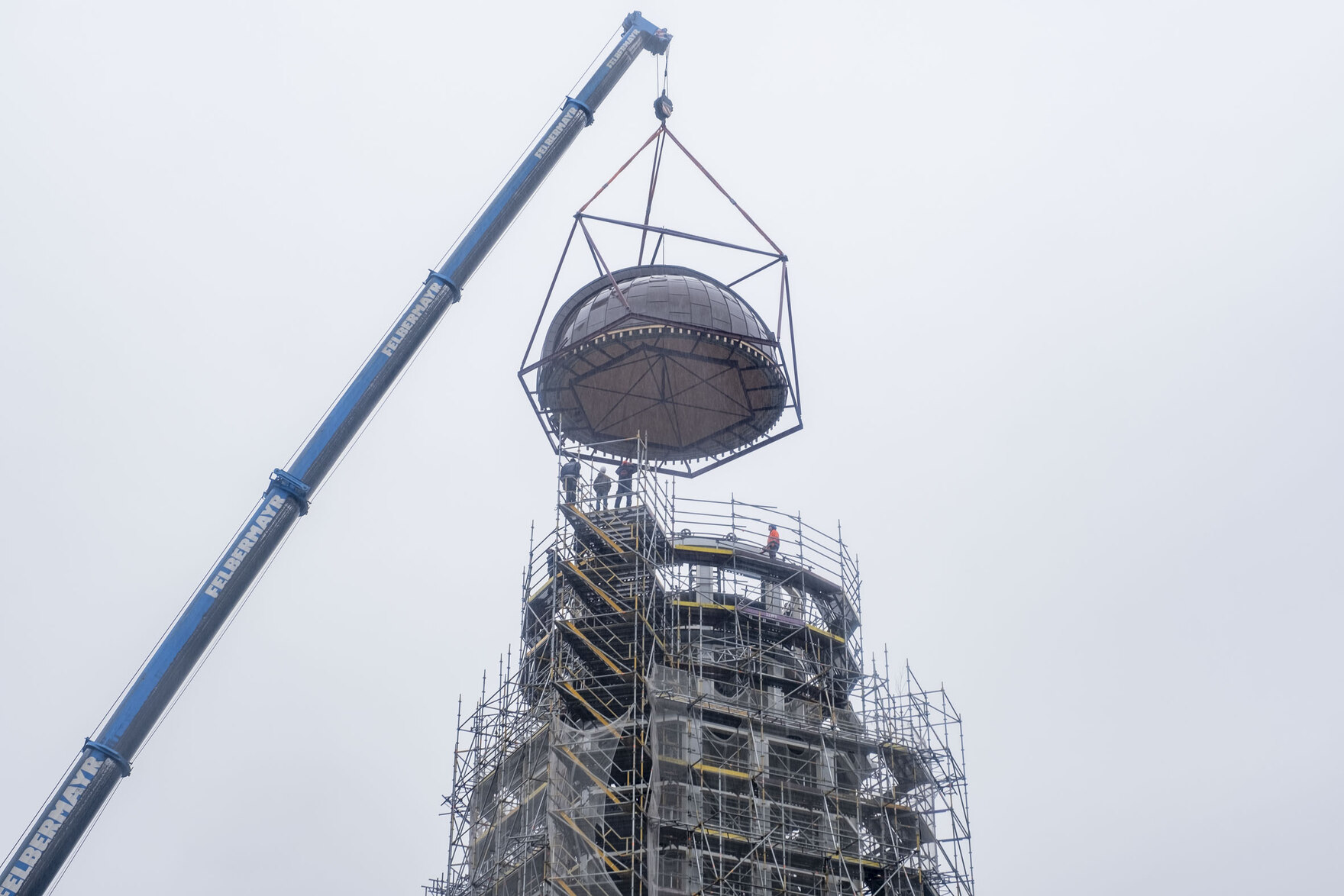 Nachtrag: Wie geplant wurde die Kuppel am 19.10.2023 montiert. Bei strömenden Regen wird die Kuppel durch einen Kran auf die sanierte Konstruktion aufgesetzt und Arbeiter montieren sie von aufgestellten Gerüsten aus.