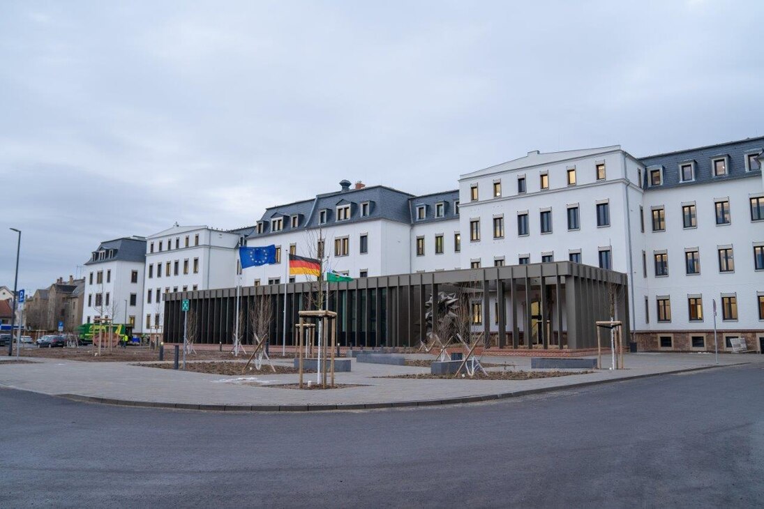 Das sanierte Kasernengebäude hat eine weiße, verputzte Fassade und graue Dachschindeln. Vor dem Haupteingang befinden sich Fahnenmasten, das Kunstwerk und daneben der neu gebaute Beratungssaal.