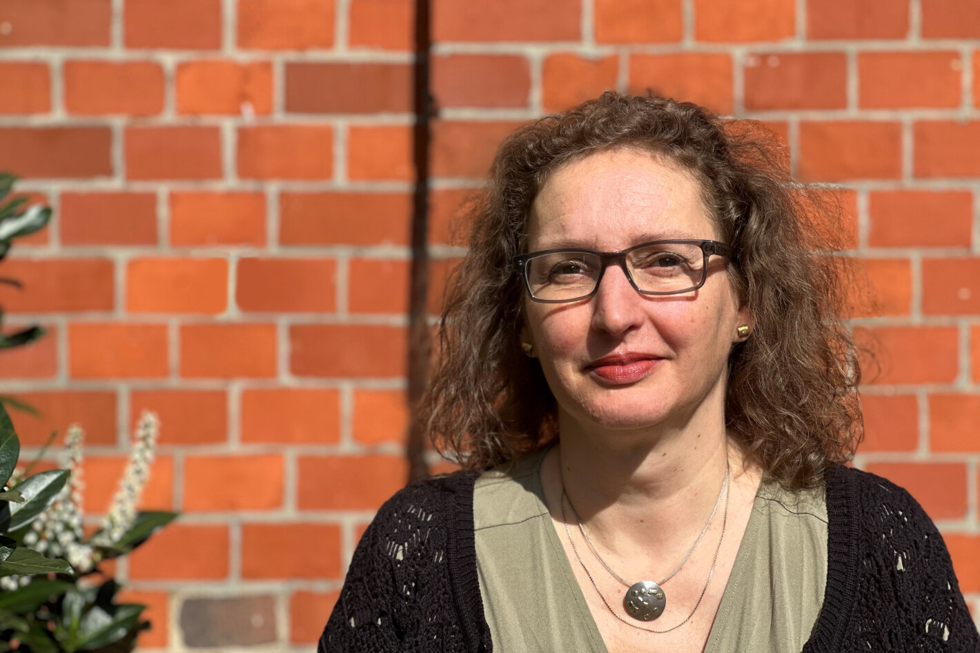 Dunkelhaarige Frau mit gelocken Haaren und Brille vor einer Ziegelsteinmauer
