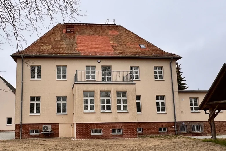 Das sanierte Gebäude hat einen helle ockerfarbene Hülle und im unteren Bereich ist Zigelmauerwerk sichtbar. Diese passen zu den roten Dachschindeln.