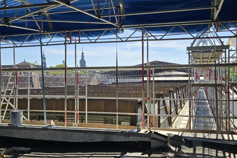 Niedrige Gerüste umringen eine sägezahnförmige Dachkonstruktion, während im Hintergrund bereits ein Teil der Dachkonstruktion mit einem flachen Wetterschutzdach überbaut ist.