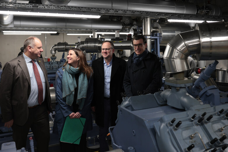 Drei Männer und eine Frau stehen in einem Technikraum mit jeweils einer Wärmepumpe vor und hinter ihnen