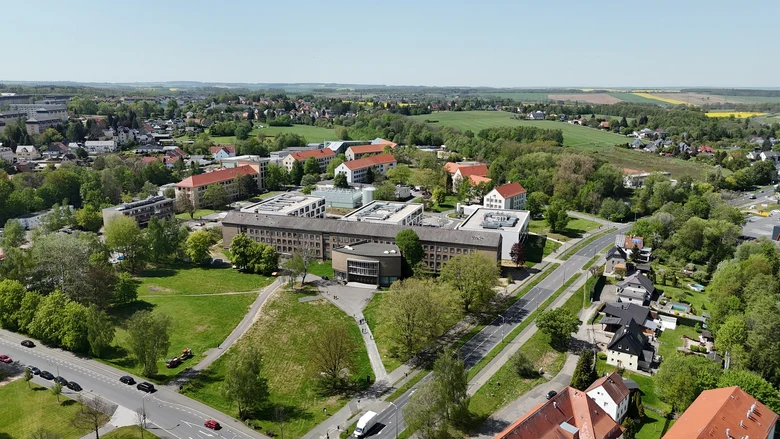 Luftbild mit einem langgezogenen, dreigeschossigen Altbau mittig im Areal und dahinter drei identische helle Gebäude senkrecht zum Altbau angeordnet