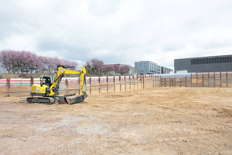breite Baugrube längs zum Hang mit holzgesicherten Grubenwänden und einem gelben Bagger sowie Kirschbäume an einer langen Grubenseite und weitere graue Gebäude an einer der kurzen Seiten
