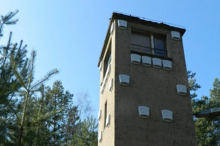 alter quadratischer Postenturm vor blauem Himmel und im Wald, der weiße Kästen an der Fassade hat