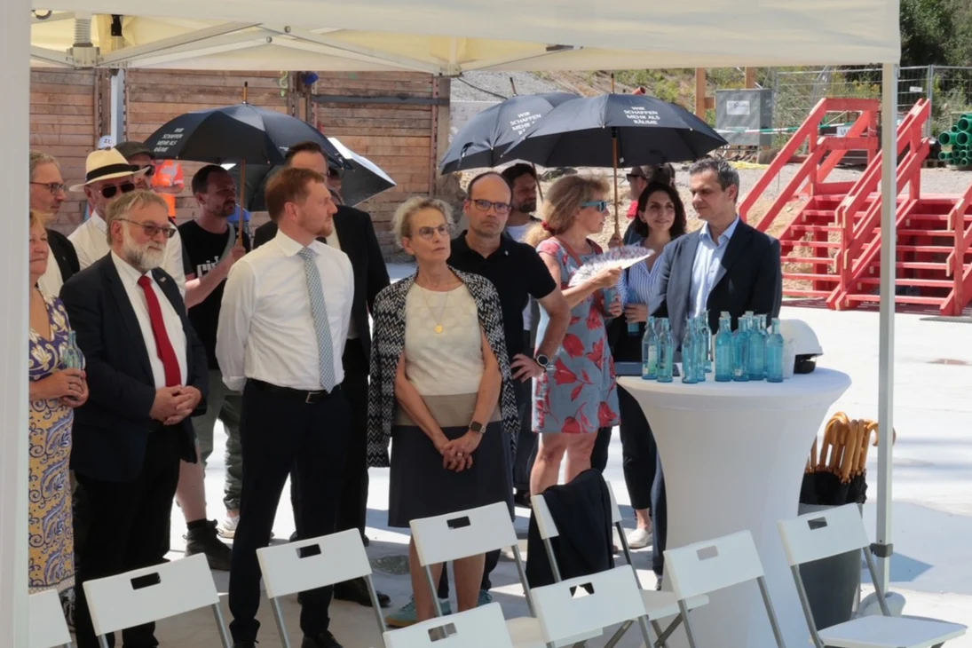 Einige Menschen stehen unter einem Pavillon während ein andere Teil der Menge schwarze Regenschirme gehen den Sonneneinfall nutzt. Vor Ihnen sind Stühle und Stehtische aufgereiht.