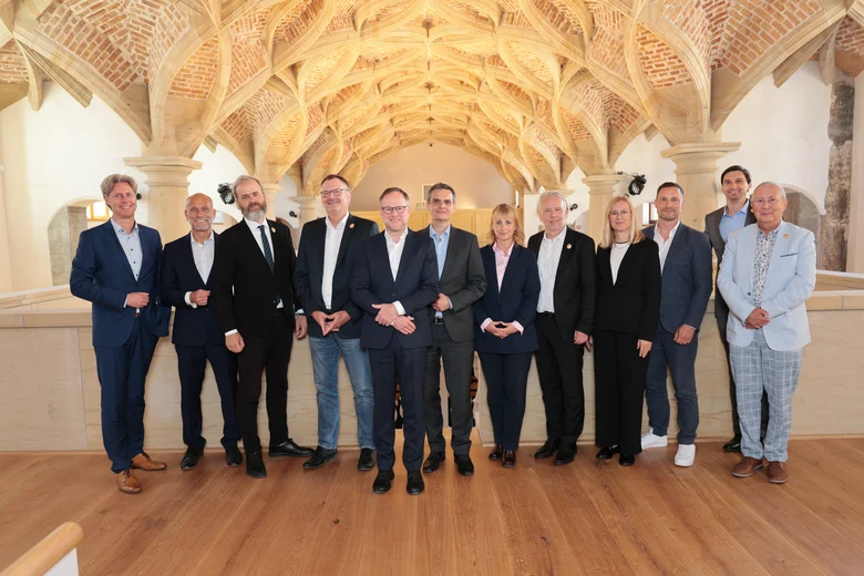 eine Gruppe von zehn Männern und zwei Frauen, die meist im Anzug gekleidet sind und auf Dielen für einer Sandsteinbrüstung unter einem Schlingenrippengewölbe stehen