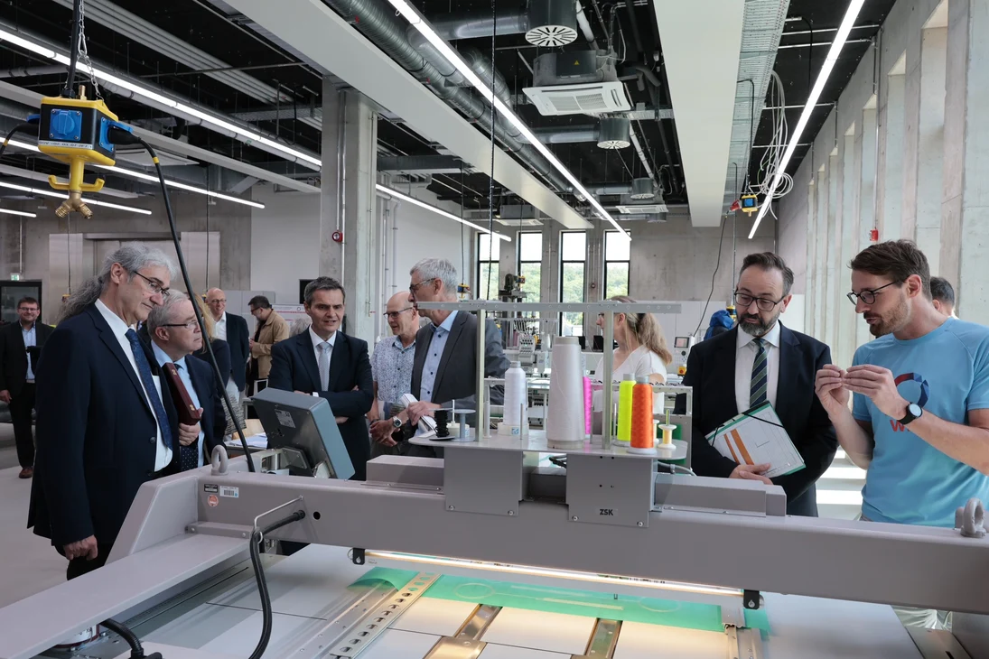 Eine Gruppe von Männern steht in einer Halle vor einer tischähnlichen Maschine, die ein Muster näht