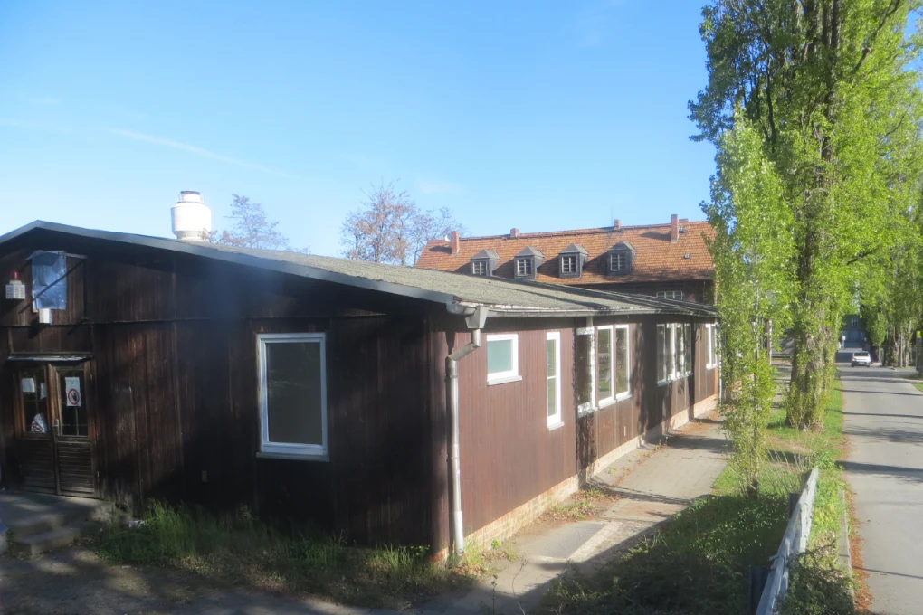 Eine eingeschossige Baracke mit dunkelbraunen Holzwänden und weiß umrahmten Fenstern an einer kleinen asphaltierten Durchfahrtsstraße, die von Bäumen gesäumt ist.