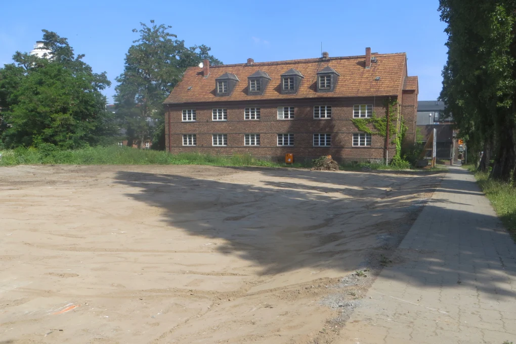 Unbebaute Fläche blanker Erde vor einem zweigeschossigen Ziegelbau mit Spitzdach aus roten Schindeln und neben einem von Bäumen gesäumten Fußweg