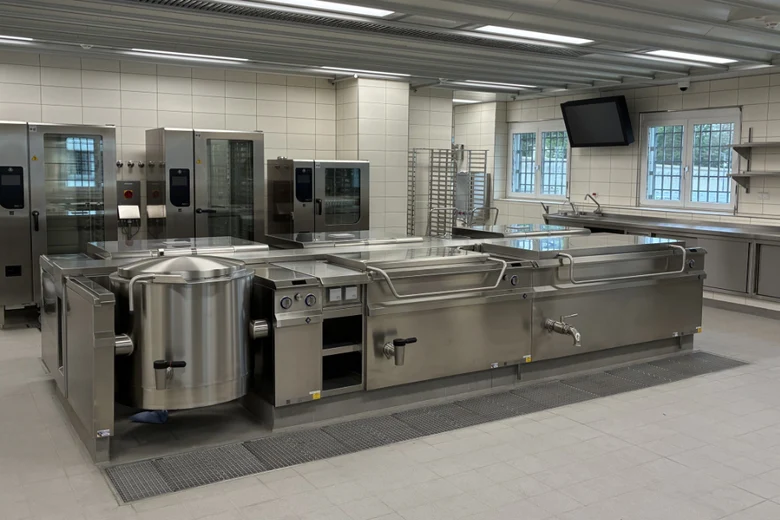 Die Großküche ist am Boden und den Wänden mit hellen, grauen Fliesen versehen. Verschiedene Arbeitsflächen wie Kochplatten, Grills, Waschbecken, Ablageflächen und Ähnlichem sind aus Edelstahl.