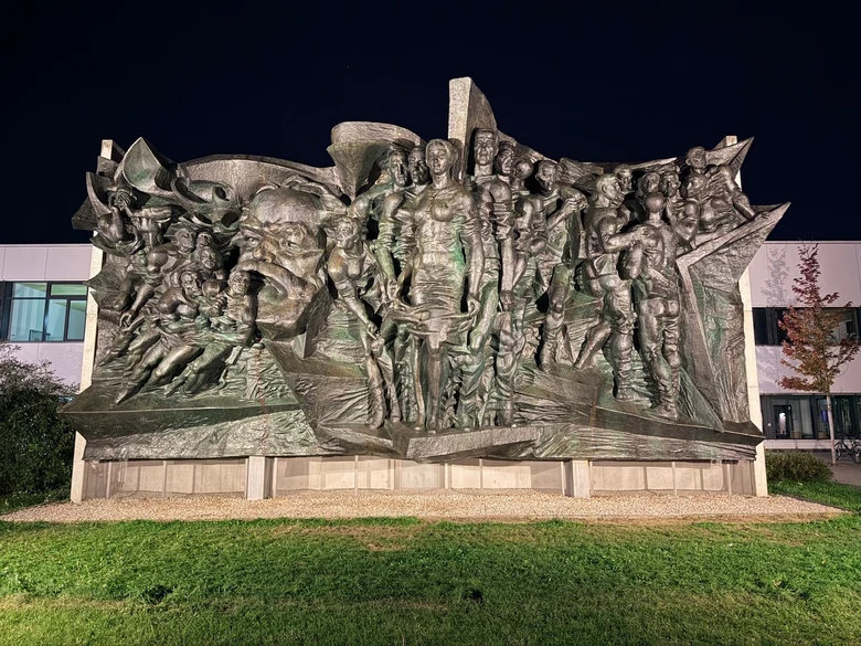 Ein großes Bronze-Relief von mehreren Personen und einem Karl-Marx-Kopf wird frontal in der Nacht angeleuchtet. An einigen Stellen zeigt sich grüne Patina. Das Relief steht auf einer Wiese vor einem Gebäude.