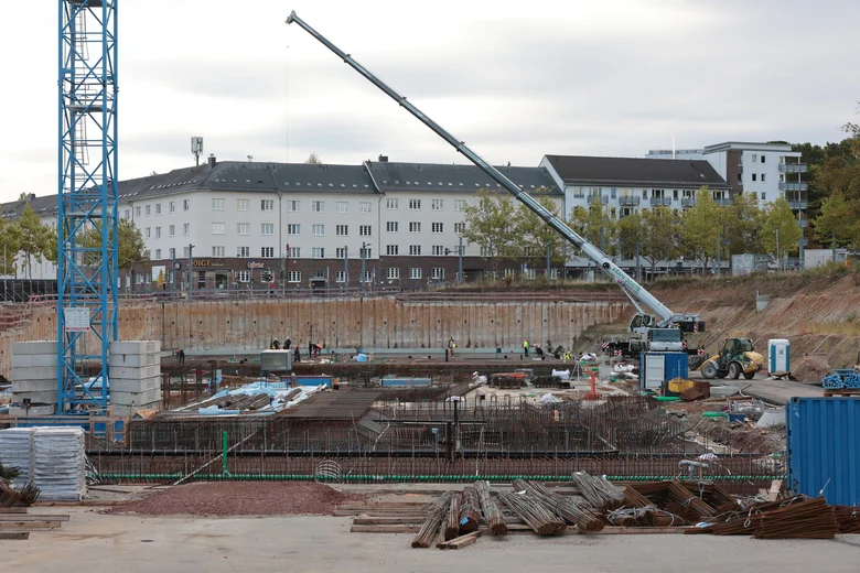 In einer großen Baugrube mit teilweise Bohrpfahlsicherung und Böschung als Baugrubensicherung befindet sich bereits eine riesige Betonplatte. Darauf liegen viele verschiedene Varianten von Bewehrungsstahl sowie andere Baumaterialien. Außerdem stehen darin