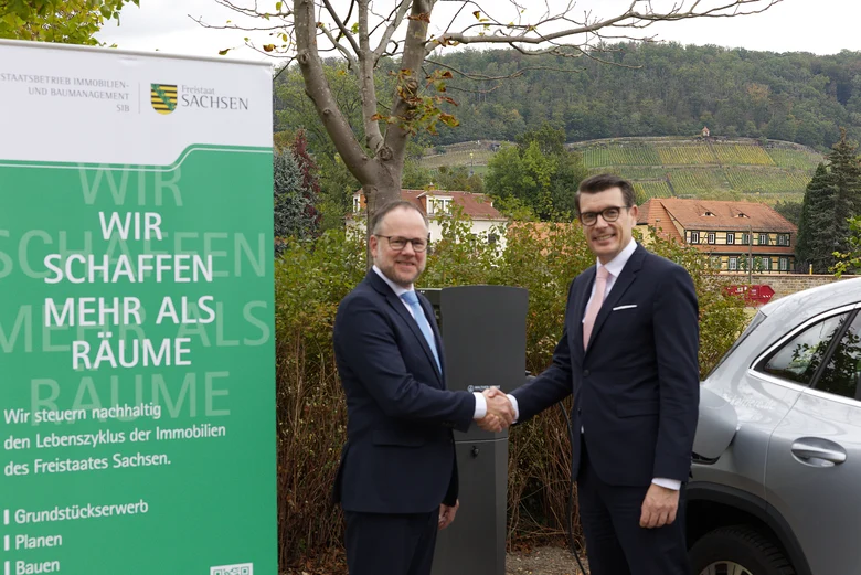 Zwei Männer in Anzügen stehen vor einer dunkelgrauen Wallbox und schütteln sich die Hand. Daneben steht ein Auto, dass an der Wallbox mittels Kabel angeschlossen ist. Dahinter befindet sich eine Hecke und noch weiter hinten ein Weinberg mit Fachwerkhäuser