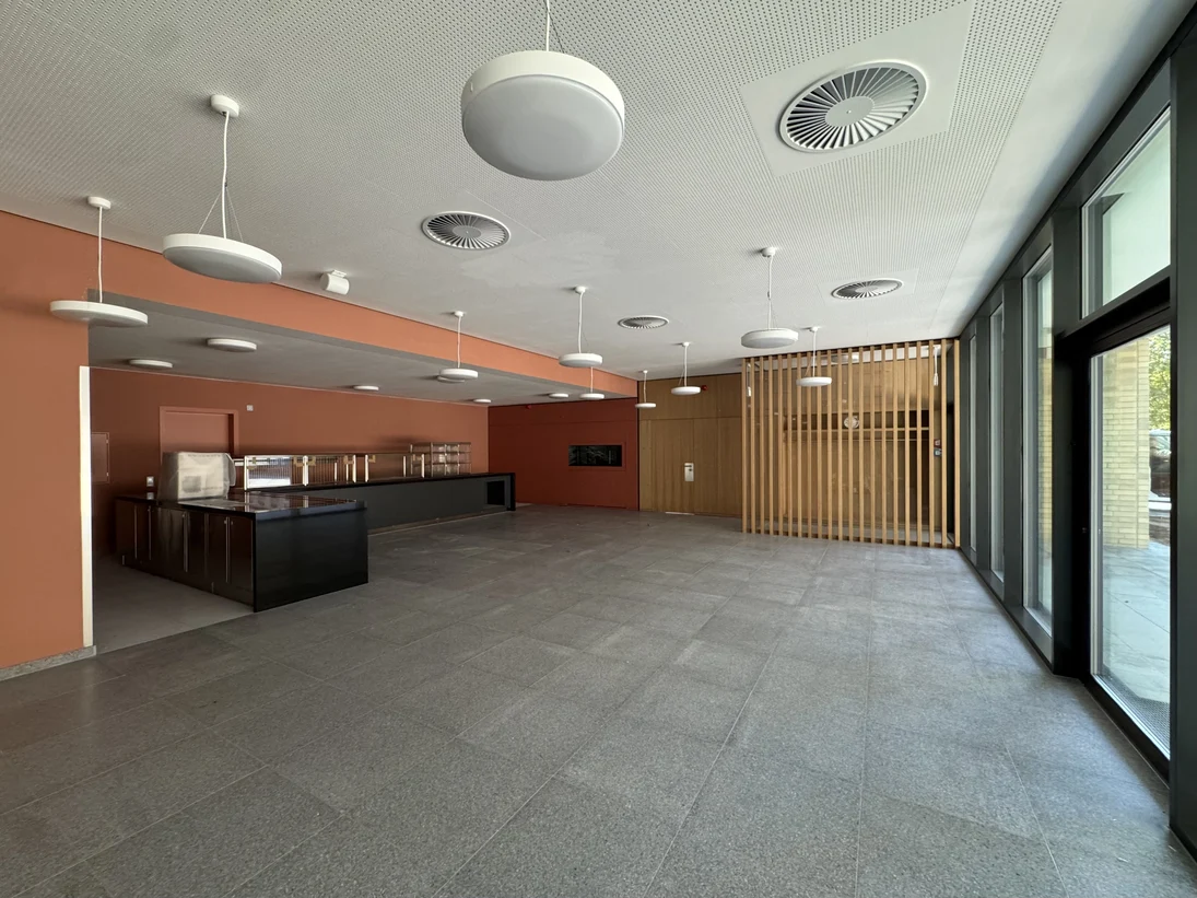 Der Speisesaal hat auf dem Boden graue Fliesen, rote Wände, teilweise mit Holzelementen, eine helle, weiße Decke und eine Glasfassade mit großen Fenstern. Eine dunkle Theke ist in der Serviceecke untergebracht.