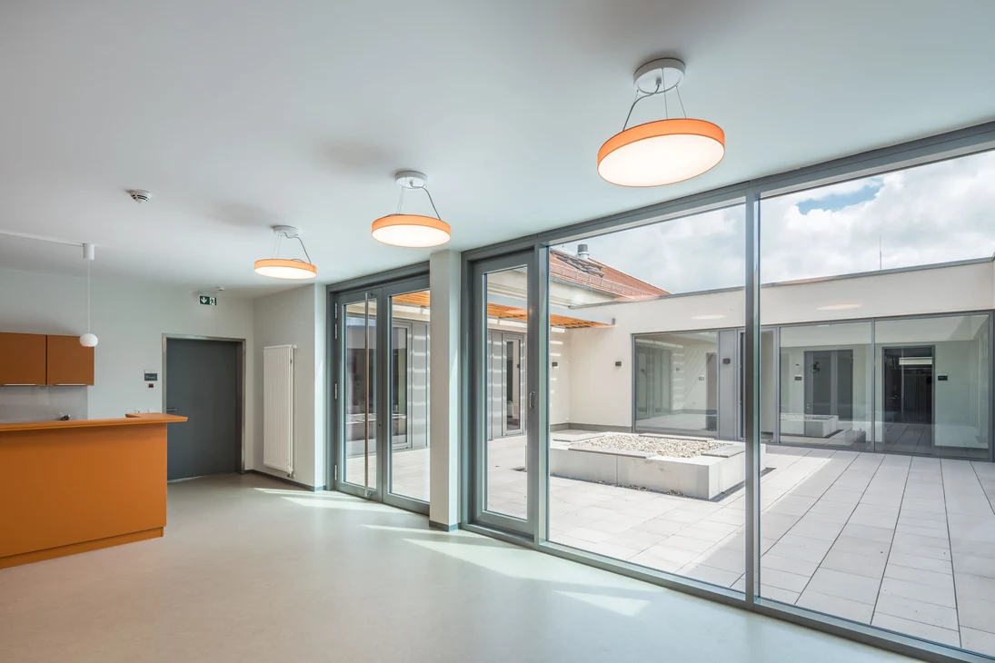 Moderner Empfangsbereich mit orangefarbenem Tresen, großen Glasfronten zu einem kleinen Innenhof und drei orangefarbenen Pendelleuchten.