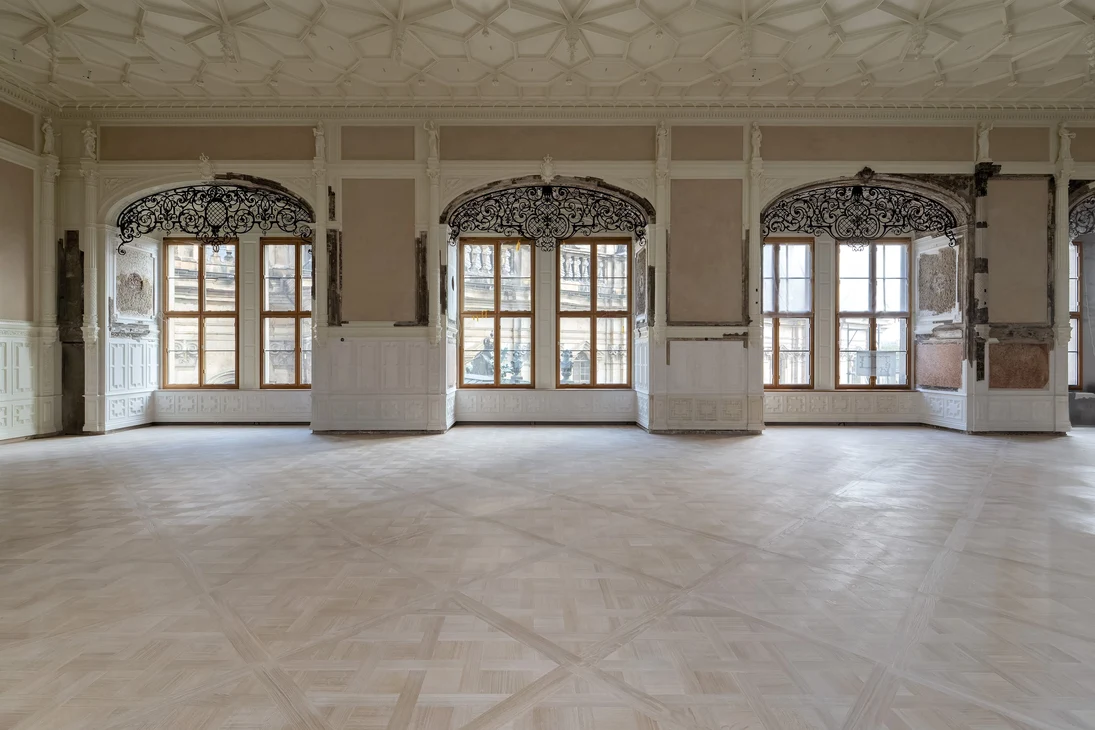 Leerer Saal, mit Parkettboden, hohen Fenstern und dekorativen  Stuckelementen an Wänden und Decke.