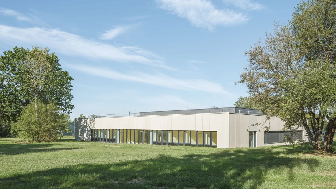 Moderner, eingeschossiger Bau mit ockerfarbener Fassade, auf grüner Wiese, von Bäumen eingerahmt und unter blauem Himmel.