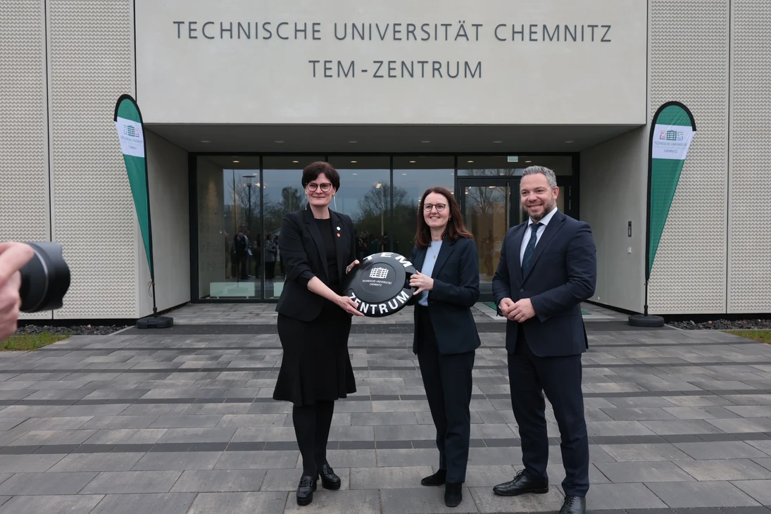Zwei Frauen halten einen überdimensionierten Transponder mit einem Mann daneben und stehen vor dem Eingang eines Gebäudes, der von Fahnen flankiert ist.