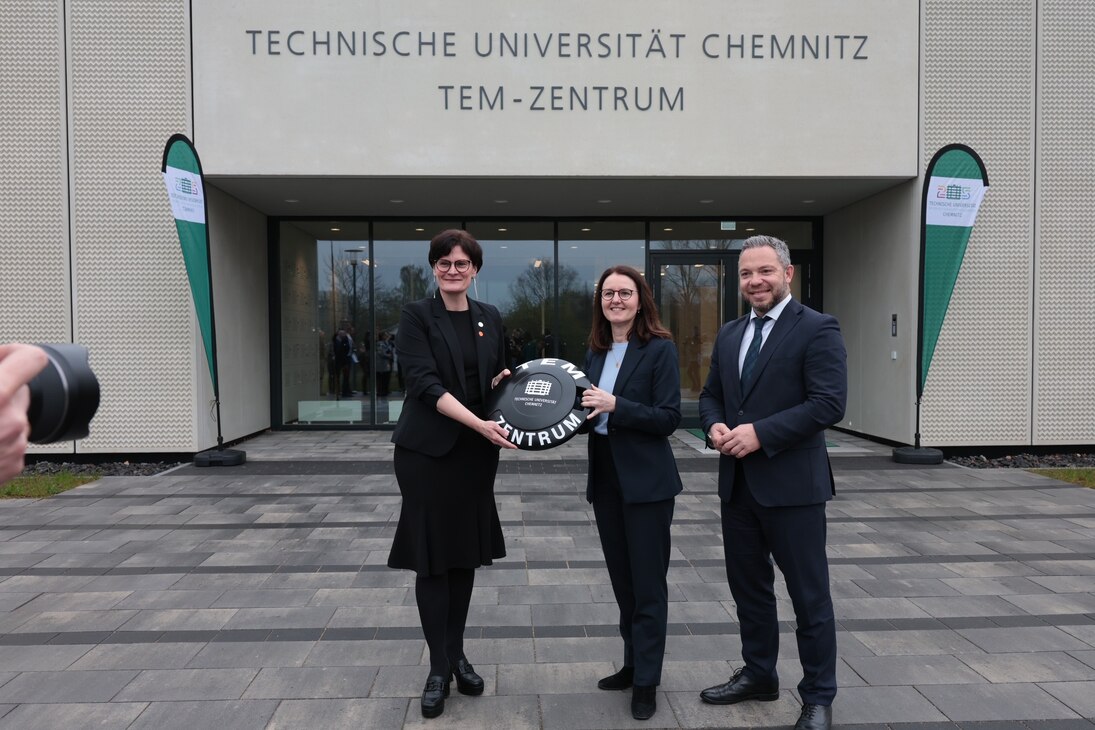 Zwei Frauen halten einen überdimensionierten Transponder mit einem Mann daneben und stehen vor dem Eingang eines Gebäudes, der von Fahnen flankiert ist.
