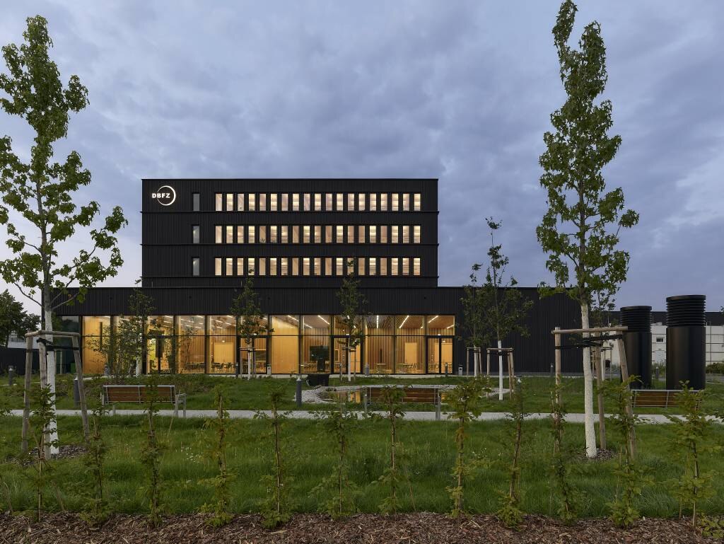 Das Gebäude hat eine dunkle Holzfassade. Im Inneren ist das Foyer aus heller Holzverkleidung erkennbar.