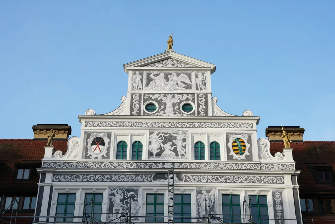 Historische, reich verzierte Giebelfassade mit  Ornamenten, Wappen, Figurenreliefs und mehreren Fenstern vor blauem Himmel.