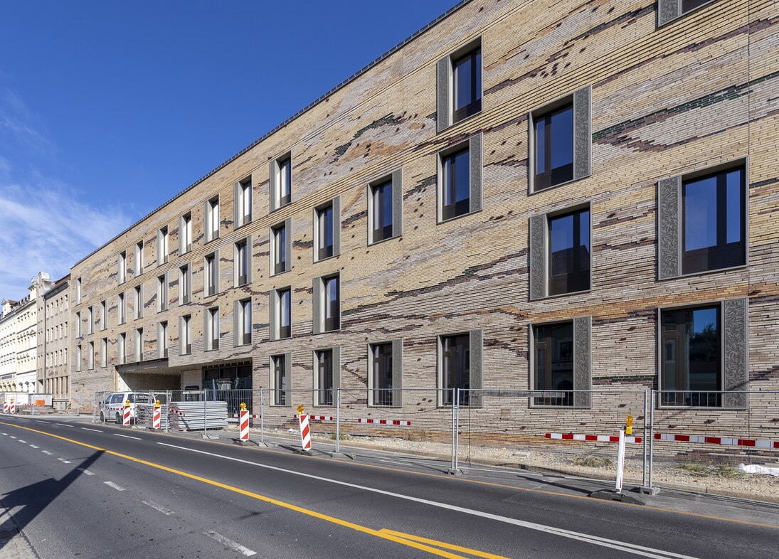 Zu sehen ist die Fassade des neuen Senckenberg-Campus Görlitz von der Bahnhofstraße Ecke Jakobstraße aus. Sie ist mit lehmfarbenen Klinkern verkleidet, die optisch durch Farbunterschiede einen Querschnitt durch Sedimentgestein darstellen. (Ansicht 1)